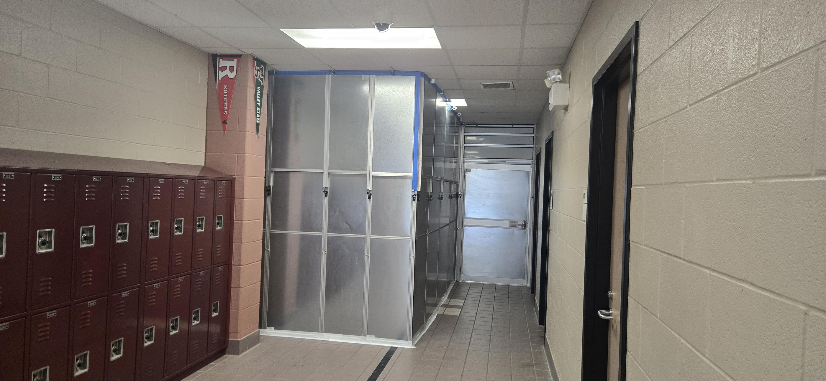 Metal-faced temporary containment wall and emergency exit door installed beside lockers in a middle school hallway.