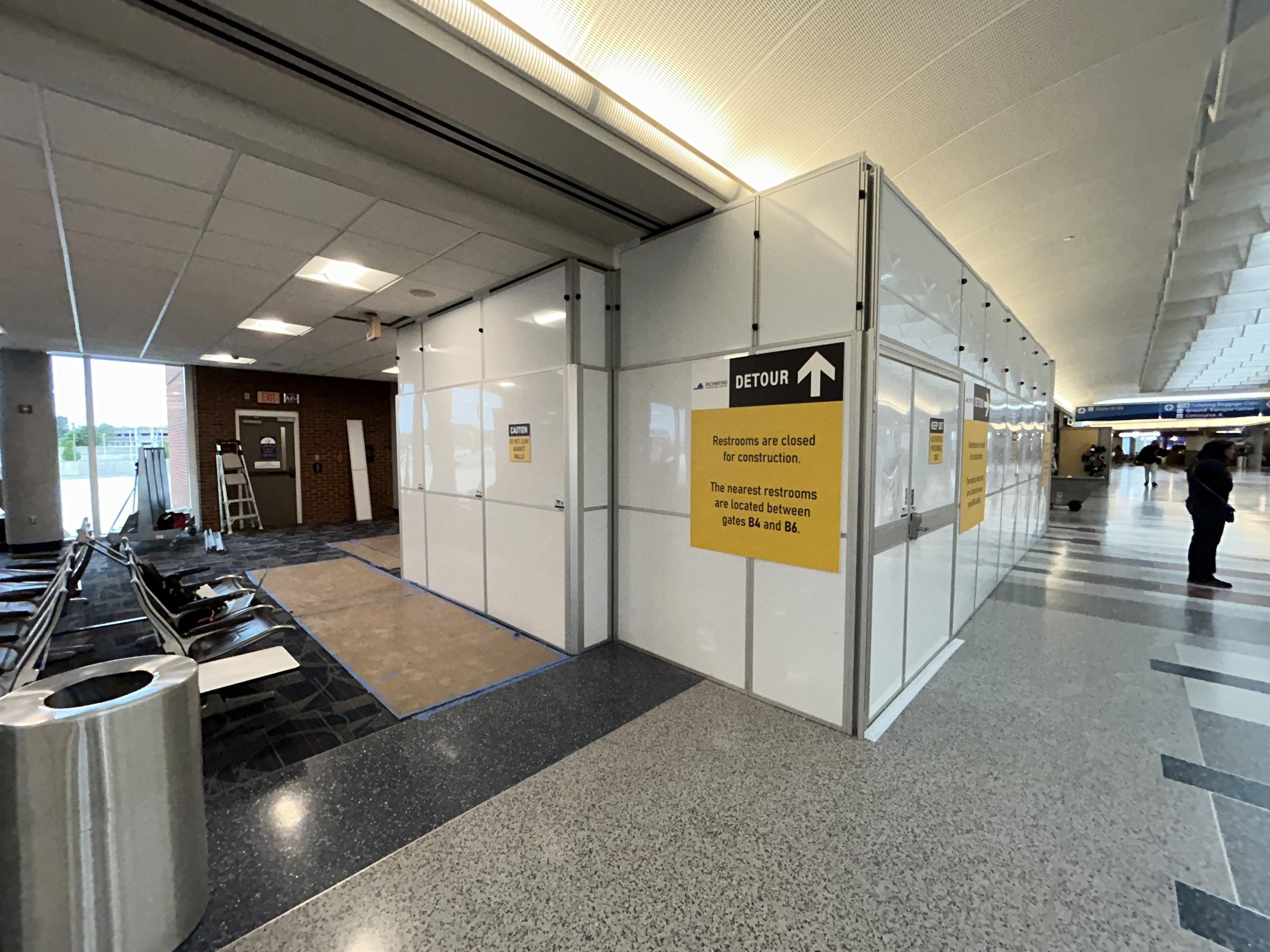 Large temporary wall enclosure with access doors and restroom detour signs separating construction from the airport concourse.