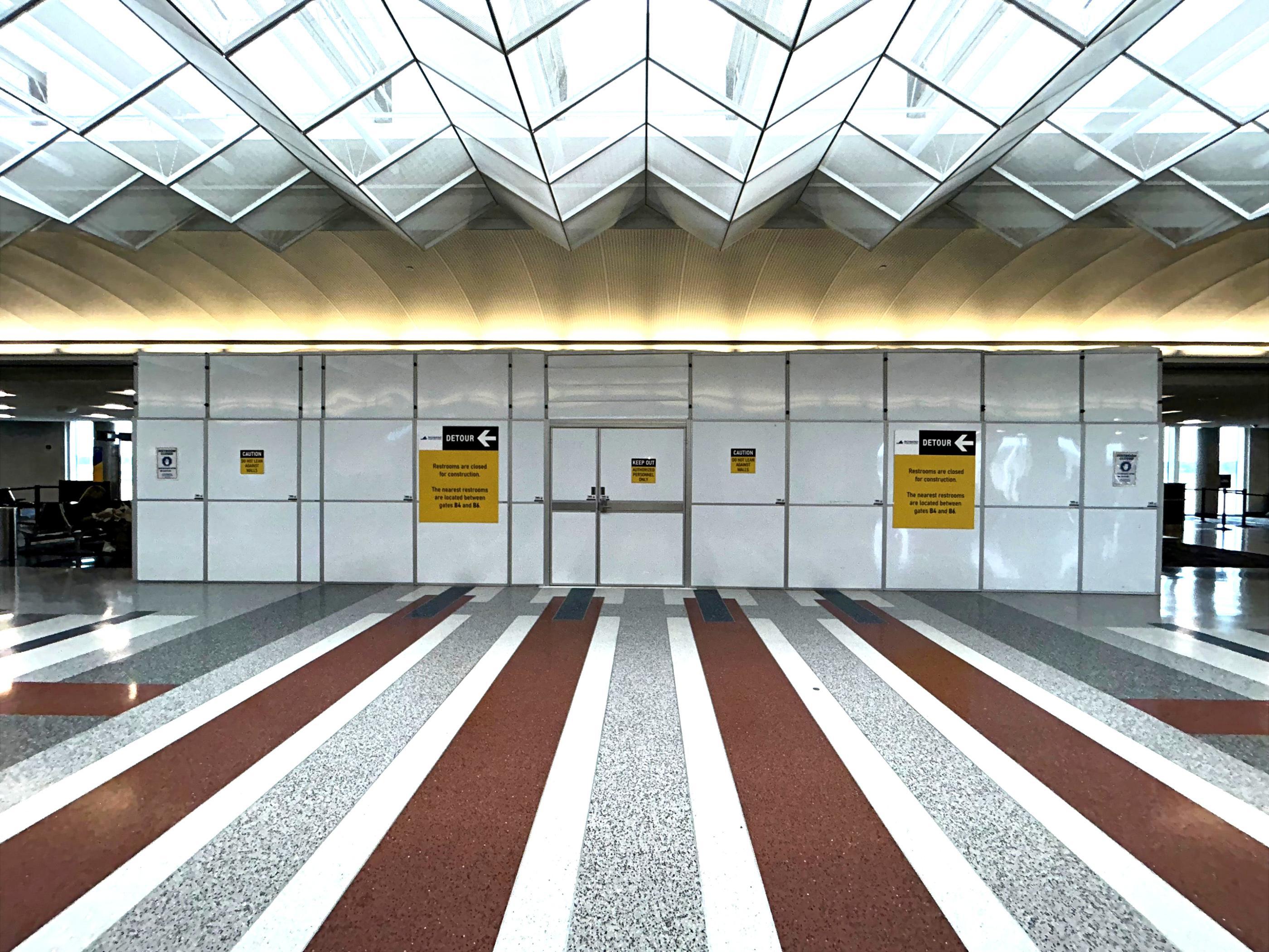 Wide temporary containment wall with double access doors and restroom detour signs inside an airport concourse.